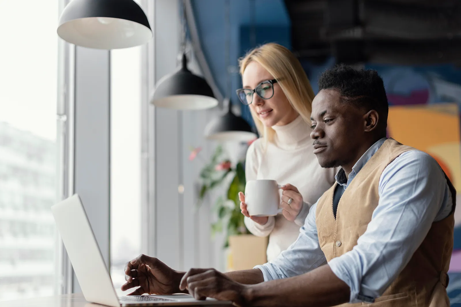 Coworkers reviewing laptop screen together at window desk with pendant lighting